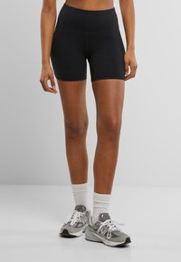 Shorts de sport noirs avec une taille haute. Tissu lisse et extensible. Portés avec des baskets grises et des chaussettes blanches montantes.