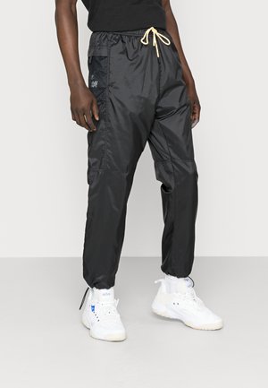 Persona que lleva pantalones deportivos negros de nylon con detalle de bolsillo de malla y cintura con cordón, combinados con zapatillas blancas y calcetines.