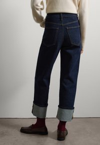 Jeans de mezclilla azul oscuro de cintura alta con puños enrollados, cierre de botón, dos bolsillos traseros y tejido de color azul más claro en los puños.