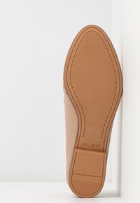 Semelle d'une chaussure beige avec une semelle en caoutchouc texturée et le nom de la marque ALDO au talon, debout contre un mur d'angle blanc.