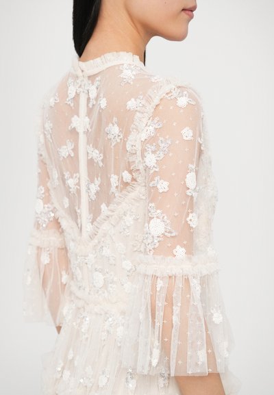 Rochie crem transparentă cu broderie florală, manșete cu volane și paiete. Prezintă un decolteu cu volane și detalii asemănătoare dantelii pe spate.