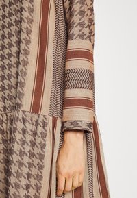 Hnědá látka s vzorem houndstooth a chevron, obsahující pruhy různých šířek; texturovaný materiál; detail tříčtvrtečního rukávu.