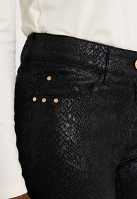 Pantalon noir texturé avec un motif subtil de peau de serpent, équipé d'accents métalliques dorés et de trois clous décoratifs sur la poche avant.