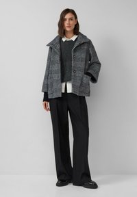 Szary żakiet w kratę z dużym kołnierzem, czarnymi guzikami, noszony na ciemnym swetrze i białej koszuli, zestawiony z luźnymi czarnymi spodniami i butami.