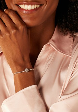 Zilveren armband met een gestructureerde slangenketting en een rond, met kristallen versierd bedeltje om een pols. De huidtint is rijk en warm; het shirt is lichtroze.