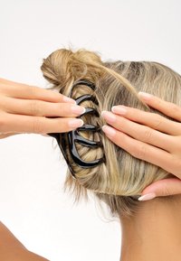 Capelli biondi raccolti in un bun fissato da un fermaglio in plastica nera, con mani che sistemano il fermaglio e unghie con manicure francese visibili.