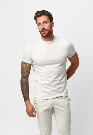 Mand med skæg og detaljeret fuldærmet tatovering iført tætsiddende hvid T-shirt og cremefarvede bukser, stående mod en ensfarvet baggrund.