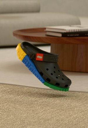 Schwarzer Clog-Schuh mit bunten Lego-ähnlichen Sohlen aus gelben, blauen und grünen Teilen, drinnen in der Nähe eines Holztisches ausgestellt.