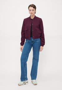 Burgundy bomber jacket with snap buttons, featuring two front pockets, paired with blue straight-leg jeans and light-colored sneakers.