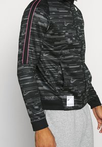 Svart zip-up-jacka med grafiskt mönster, som har rosa detaljer på axlarna. Tillverkad av slätt tyg med elastiska muddarna och nederkanten.