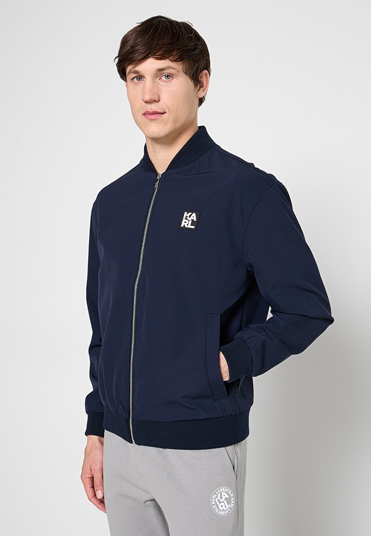Joven con pelo corto y castaño que lleva una chaqueta azul marino con cremallera y el logo "KARL" y pantalones deportivos gris claro con un logo circular.