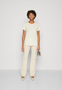 Camiseta crema con logo "Guess" de lentejuelas, combinada con pantalones claros estampados. Los accesorios incluyen un bolso plateado y zapatos beige.