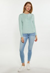 Lichtblauwe cardigan van zacht materiaal, met een ronde hals, knoopsluiting en lange mouwen, gecombineerd met strakke blauwe jeans en witte sneakers.