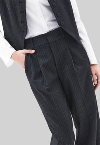 Donkergrijze broek met fijne krijtstrepen, een platte voorkant, subtiele textuur en zakdetail. Gecombineerd met een wit overhemd met kraag en een vest.