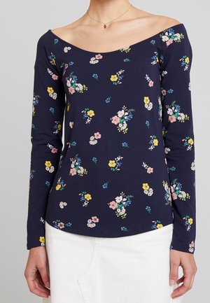 Haut manches longues bleu marine avec motif floral rose, jaune et blanc, encolure décolletée, porté avec un bas blanc et un collier doré.