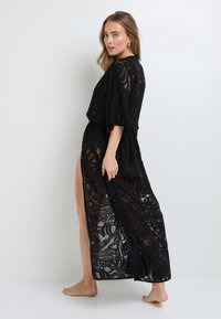 Kimono in pizzo nero con apertura frontale, maniche corte e uno spacco laterale. Motivi floreali intricati creano un aspetto trasparente e testurizzato.