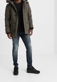 Parka aislante de color verde oliva con capucha forrada de piel, camiseta de manga larga negra, vaqueros azules desgastados y zapatillas negras.