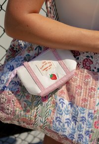 Bras tenant un petit sac blanc et rose avec un patch fraise et le texte "The Flat Lay Co. summer club" sur un tissu matelassé floral coloré.