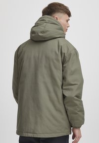 Oliven grønn polstret jakke med hette, med strukturert stoff, elastiske ermer og rett kant. Bakvisning viser enkelhet i designet.
