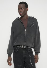 Dunkelgrauer Kapuzenpullover mit Reißverschluss und Fronttasche, verziert mit dem genieteten Buchstaben „H“. Getragen über einem braunen Oberteil, kombiniert mit abgenutzten schwarzen Jeans und einem genieteten Gürtel.