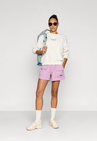 Άσπρο φούτερ με στρογγυλή λαιμόκοψη, μωβ cargo σορτς με τσέπες, κρεμ αθλητικά παπούτσια και στρογγυλά καφέ γυαλιά ηλίου. Το σακίδιο είναι ανοιχτό μπλε.