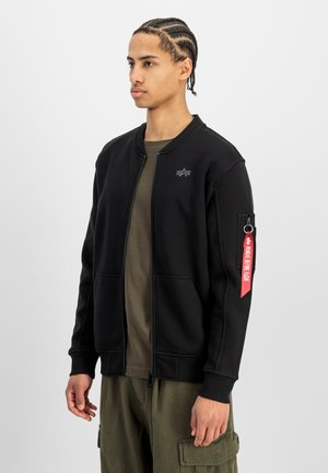 Joven con trenzas, llevando una chaqueta negra con cremallera con etiqueta roja "Remove Before Flight" en la manga y pantalones cargo verde oliva.