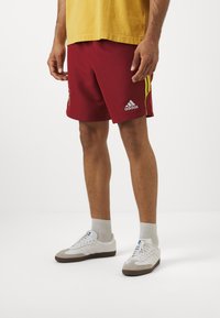 Røde idrettsshorts med gule sidestreker, Adidas-logo og et lett stoff. Brukt med hvite joggesko og grå sokker.