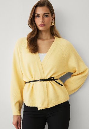 Kvinde med bølget hår iført en blød gul pakketrøje, der er spændt ind med et tyndt sort bælte, kombineret med sorte bukser og store gyldne øreringe.