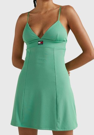 Woman wearing a fitted, ribbed, mint green mini dress with thin straps and a Tommy Jeans logo on the chest.
