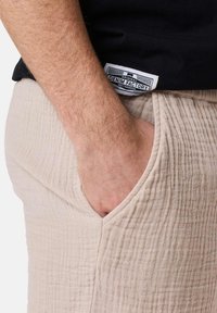Pantalons beige texturés avec une finition froissée, dotés de poches latérales et d'une étiquette cousue dans la ceinture, partiellement visible.