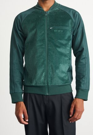 Mand iført en mørkegrøn Adidas lynlåstrøje med kordfront og glatte ærmer, sat sammen med sorte bukser, stående mod en hvid baggrund.