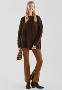 Bruine gebreide trui met kabelontwerp, gecombineerd met bruine ribfluwelen broek met zijslits, en houdt een tas met luipaardprint vast.