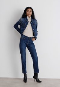 Denim takki, jossa on niittikoristeita, kerma värinen kauluspaita, siniset farkut ja mustat nilkkurit, kaikki neutraalilla taustalla.