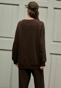 Pruun üli suur स्न sweatshirt ribastest varrukatest ja helmest, millel on minimaalne bränding ülemises seljas, koos sobivate pükstega.