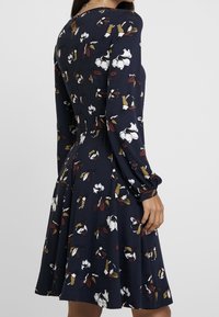 Robe marine à manches longues, motif floral en blanc, bordeaux et moutarde. Présente une taille cintrée et une jupe évasée, en tissu à texture douce.