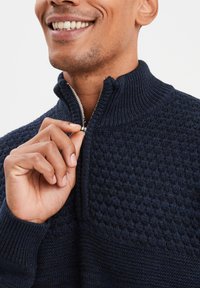 Marineblauer Reißverschluss-Pullover mit strukturiertem Muster, geripptem Kragen und glattem Strick. Nahaufnahme zeigt eine Hand, die den silbernen Reißverschlusszug hält.