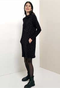 Schwarzes Hoodie-Kleid mit strukturiertem Leopardenmuster, langen Ärmeln, Fronttaschen und einem Kordelzug. Kombiniert mit schwarzen Knöchelstiefeln.
