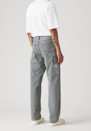 Homme portant des baskets blanches, une chemise blanche à manches courtes et un pantalon rayé vertical noir et blanc avec des poches à l'arrière et sur les côtés, debout devant un fond uni.