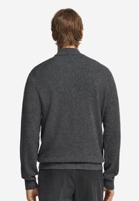 Mann mit hellbraunen Haaren von hinten gesehen, der einen dunkelgrauen Langarmpullover und schwarze Hosen trägt, vor einem schlichten weißen Hintergrund.