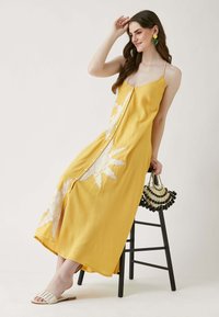 Gelbes Maxikleid mit Spaghetti-Trägern, verziert mit weißer floraler Stickerei und Knopfleiste vorn, getragen mit einer gemusterten Handtasche.