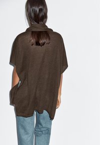 Osoba z długimi ciemnymi włosami, ubrana w luźną brązową bluzę w stylu poncho oraz jasnoniebieskie dżinsy, odwrócona tyłem przed jednolitą białą ścianą.