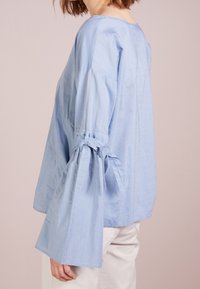 Femme portant un blouse bleu clair avec des manches larges et froncées nouées en nœuds, associée à un pantalon blanc cassé, présentée de profil devant un fond rose.