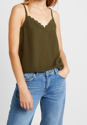 Kobieta w oliwkowej górze na ramiączkach z koronką, wpuszczonej w jasnoniebieskie dżinsy, z jedną ręką w kieszeni, na jednolitym tle.