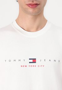 Bijela pamučna majica s printom "TOMMY JEANS" i logom u crvenoj, mornarskoj plavoj i bijeloj boji. Okrugli izrez, ležeran kroj.