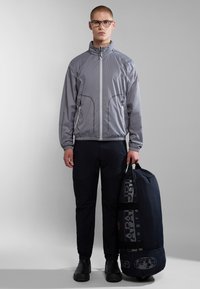 Helles graues, mit Reißverschluss versehenes Jacket mit hohem Kragen, kombiniert mit dunklen Cargo-Hosen. Hält eine marineblaue Sporttasche mit strukturiertem Design und Logo.