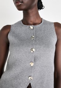 Tricou fără mâneci de culoare gri, realizat din tricot, cu decolteu rotund, având cinci butoane metalice pe față și o mică despicătură la tiv.