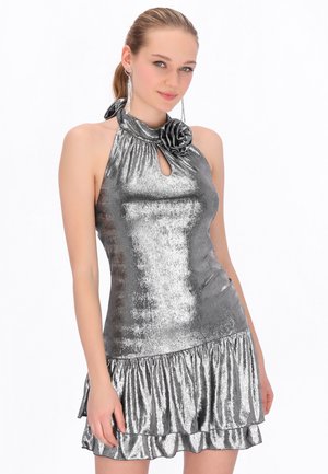 Jonge vrouw draagt een mouwloze, metalen zilveren halterjurk met een gerimpelde rok en bloemmotief bij de hals, tegen een witte achtergrond.