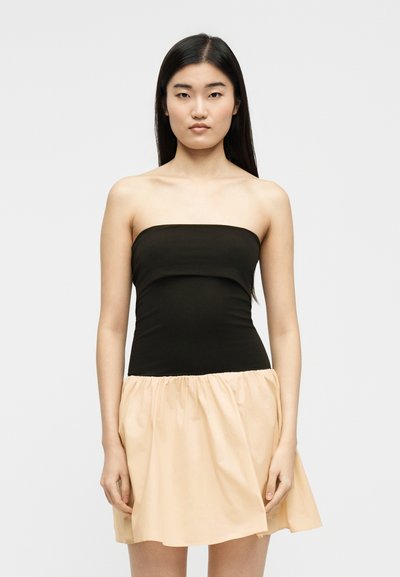 Νέα γυναίκα με μακριά σκούρα μαλλιά, φορώντας ένα μαύρο τοπ χωρίς τιράντες και μια χαλαρή μπεζ φούστα, μπροστά σε ένα απλό φωτεινό φόντο.