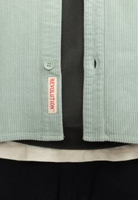 Lichtgroene corduroyjas met knoopsluiting en een label met de tekst "REVOLUTION." Draag het over een donker shirt en een crèmekleurige laag.