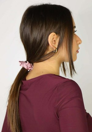 Una goma de seda rosa sujeta el cabello largo, liso y castaño. La persona lleva una camiseta ajustada color granate, visible desde el perfil derecho.